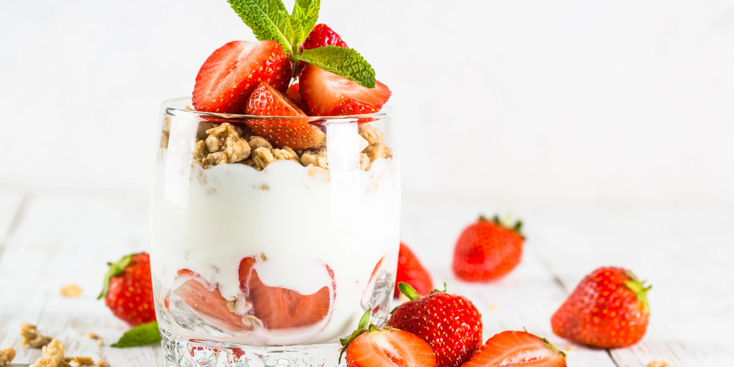 Glass full of yogurt on a bed of chopped strawberries with granola scatted on top with further scattering of granola and strawberries around the glass.